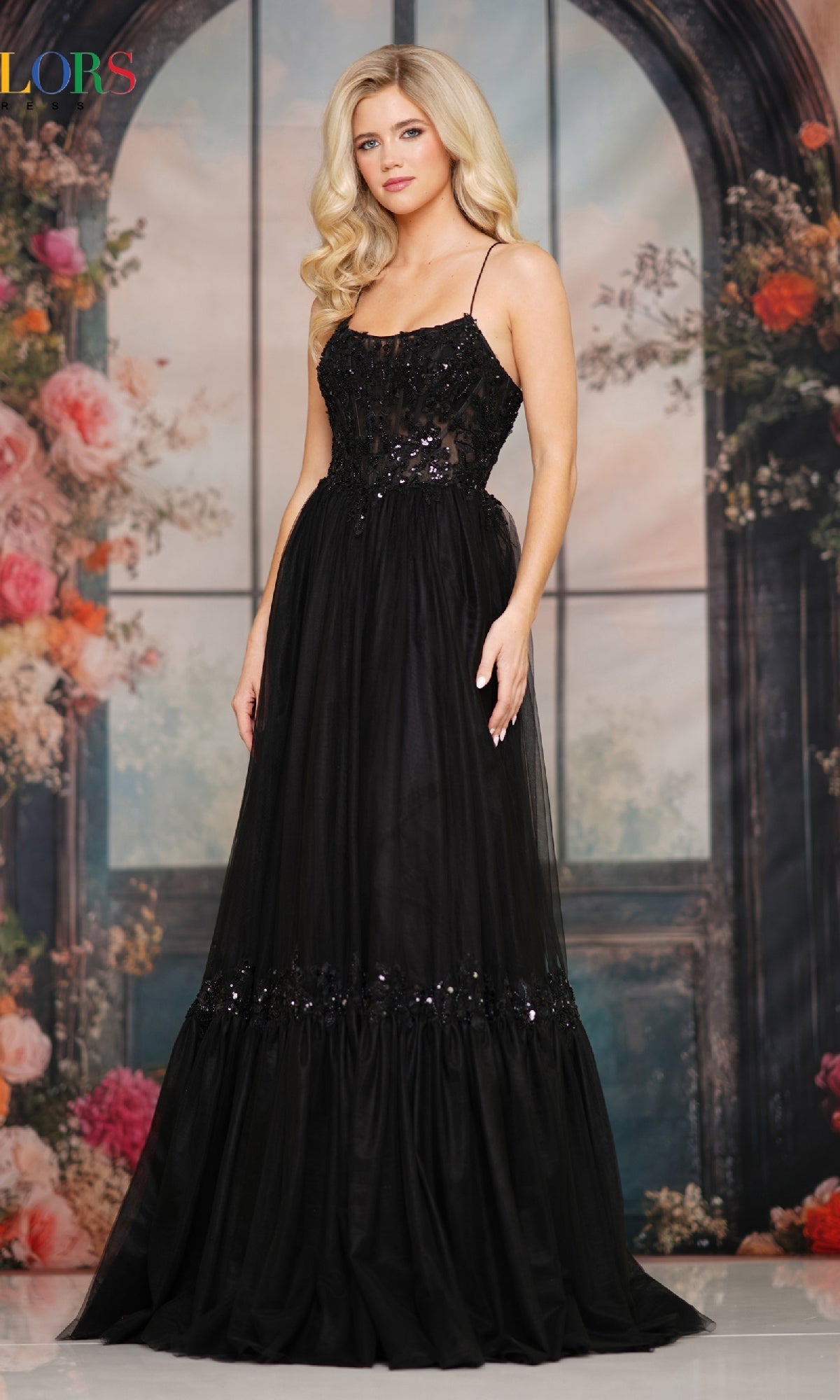 A woman in the Colors Dress 3799, a sleeveless long prom dress with sequin details and a lace-up open back, stands before a floral and arched window backdrop.